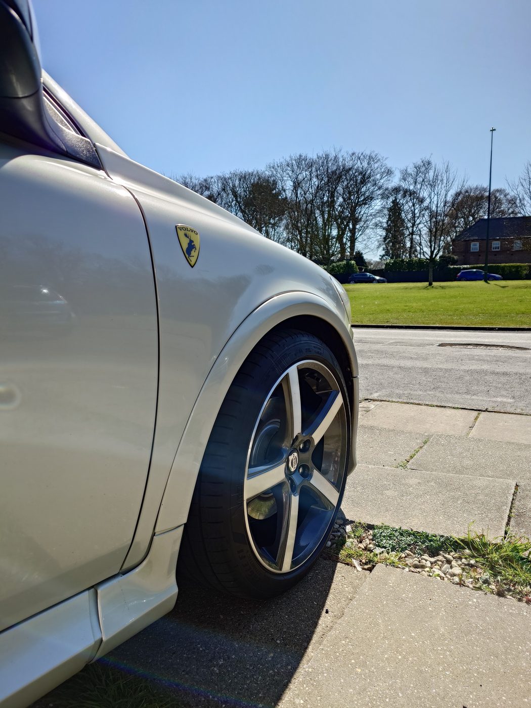 A silver sports car's front alloy wheel and tyre, parked outside on a sunny day